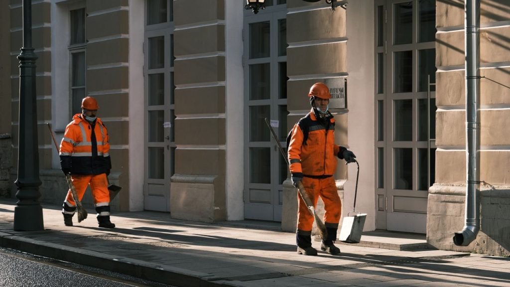 Street workers in orange safety uniforms walking along a city sidewalk in Moscow.